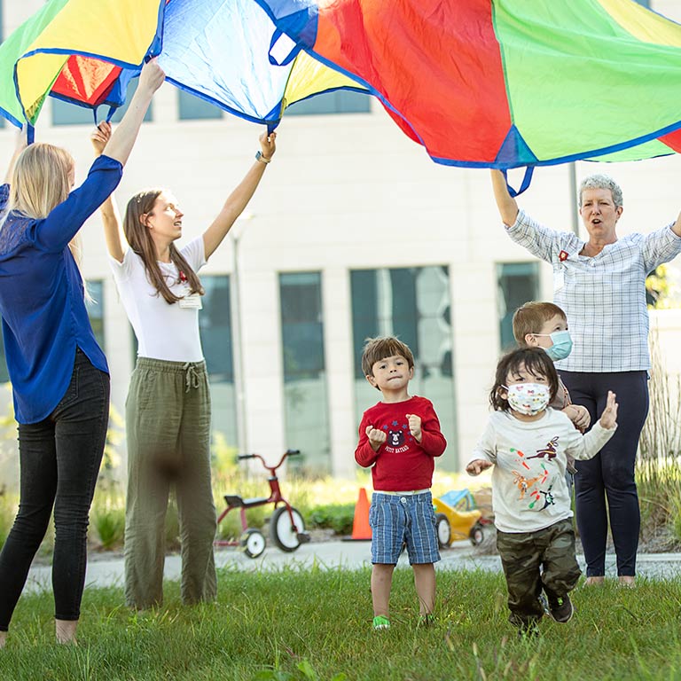Three kids run under a parachute adults hold above them in the air