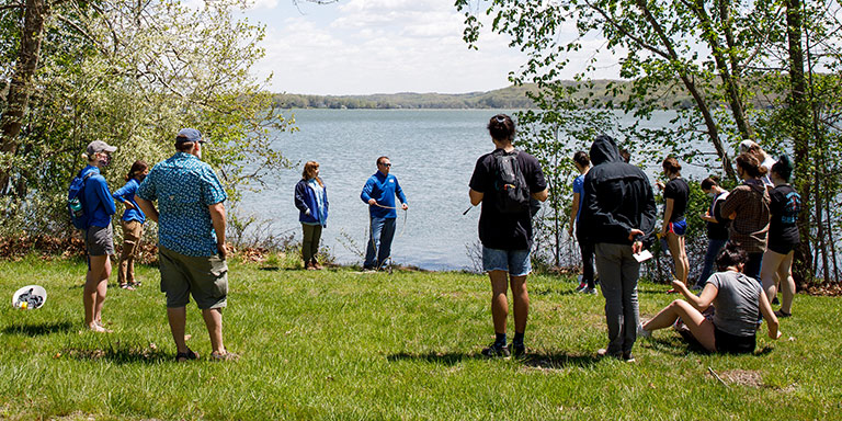 A group of people stand in a circle in the grass; a lake is visible in the background