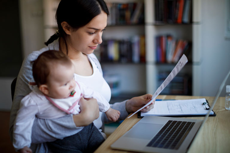 Mother holding baby while looking at paper in front of laptop