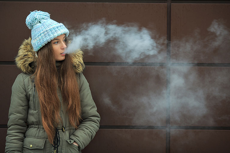 Teen girl in coat and hat breathes out vape smoke